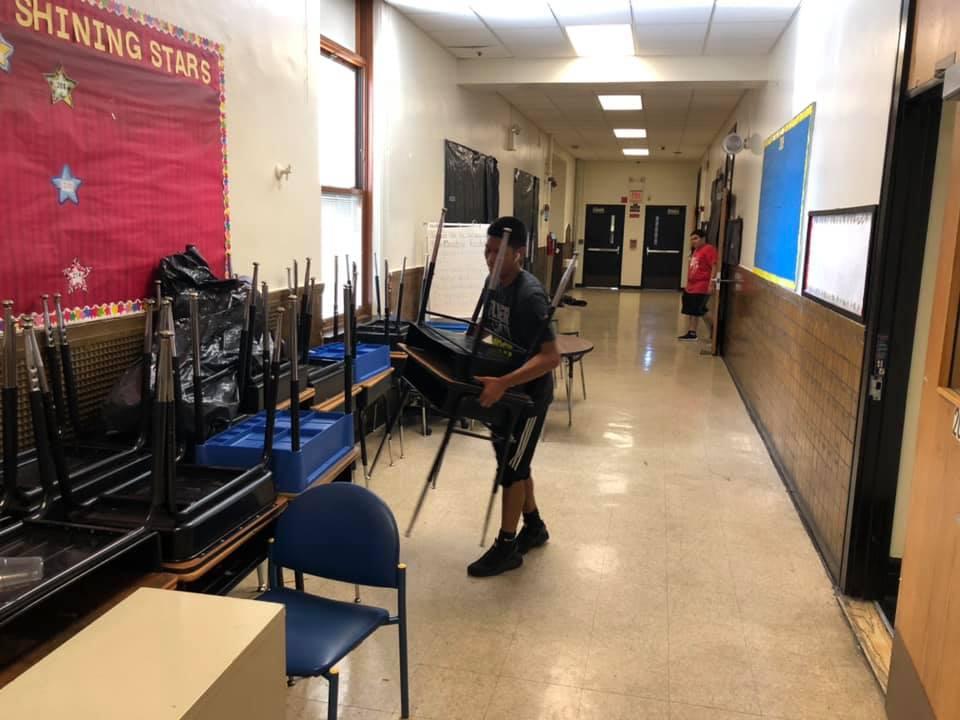 two boys taking the desks and chairs out of a classroom into the hallway